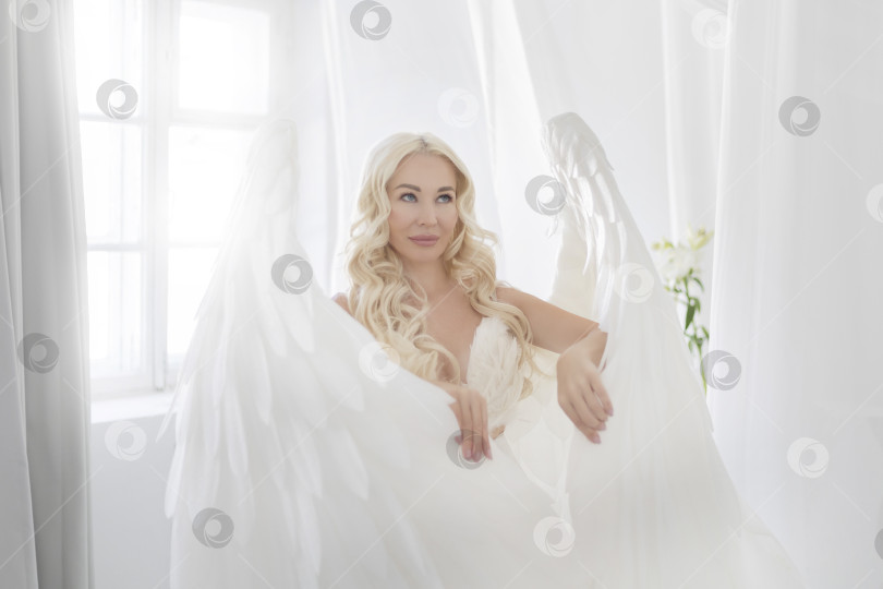 Скачать Привлекательная молодая женщина в белой тунике с большими белыми крыльями за спиной и нимбом над головой позирует, сидя в белых светящихся облаках. фотосток Ozero