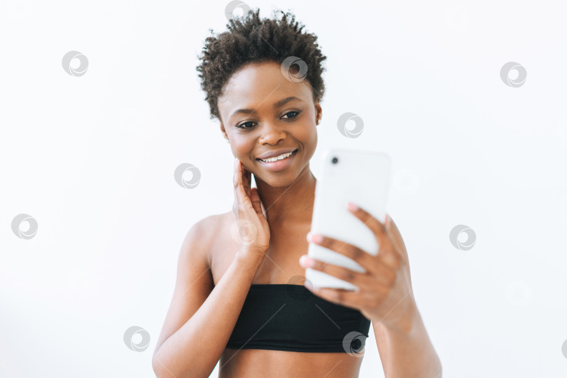 Скачать Улыбающаяся красивая молодая афроамериканка в черном топе, использующая мобильный телефон, изолированный на белом фоне фотосток Ozero