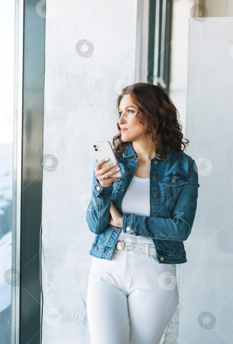 Скачать Портрет улыбающейся очаровательной молодой женщины в джинсовой куртке с мобильным телефоном у окна в современном здании фотосток Ozero