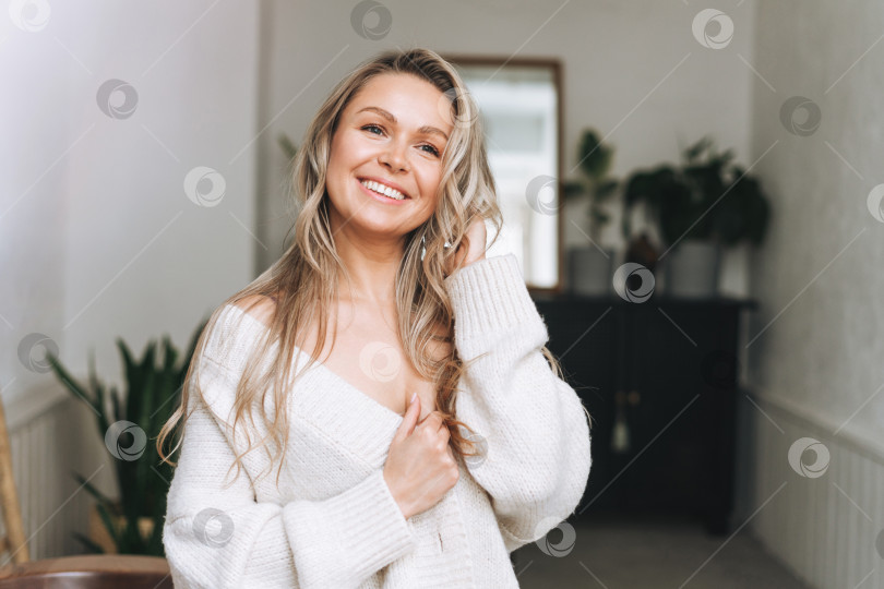 Скачать Светловолосая улыбающаяся женщина 35 лет с чистым свежим лицом и руками, длинными волосами в уютном вязаном кардигане в ярком интерьере фотосток Ozero
