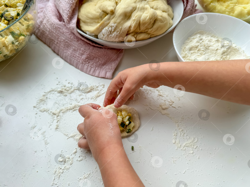 Скачать Руки ребенка замешивают тесто для приготовления пирожков на белом столе, вид сверху. фотосток Ozero