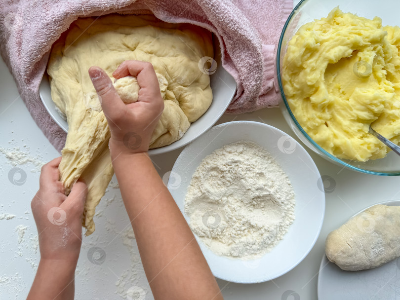 Скачать Руки ребенка замешивают тесто для приготовления пирожков на белом столе, вид сверху. фотосток Ozero