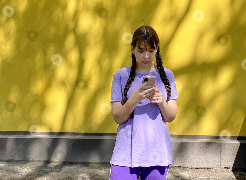 Скачать девушка в сиреневой футболке с телефоном в руках стоит у желтой стены фотосток Ozero