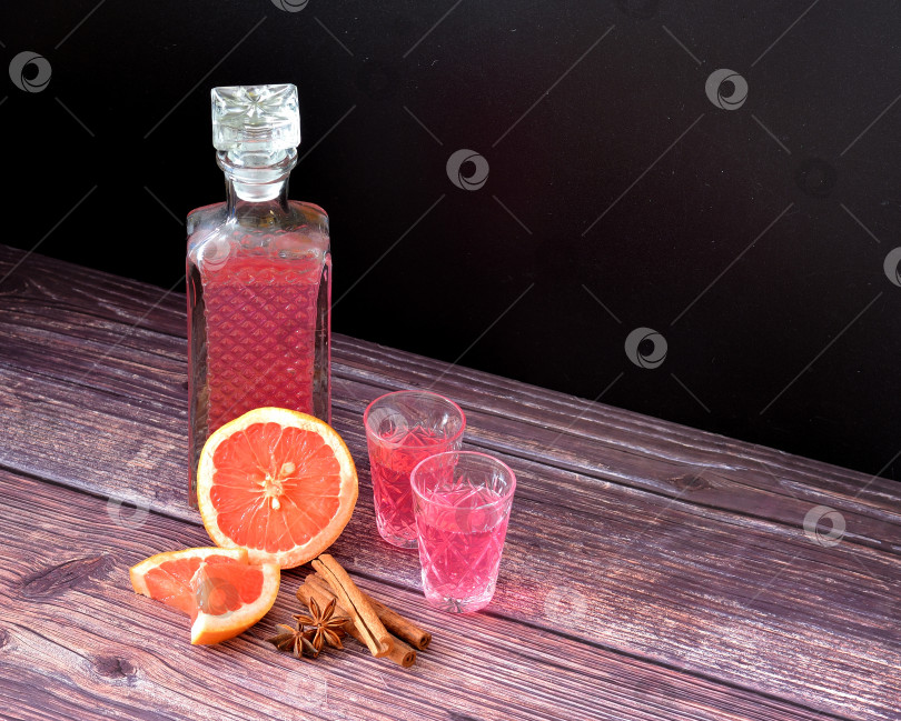 Скачать Грейпфрутовый ликер со специями на деревянном столе, крепкий домашний алкоголь в бутылке и две рюмки с корицей, анисом и кусочками спелых цитрусовых. фотосток Ozero