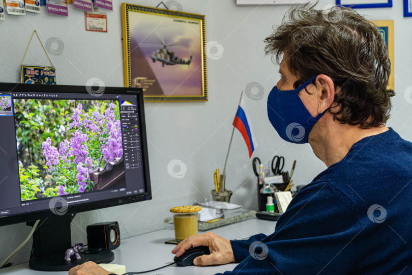 Скачать Взрослый мужчина в защитной маске на лице обрабатывает фотографии в офисе на компьютере. Размытый фон. Выборочный фокус. Коронавирус Covid-19, пандемия. Карантин. фотосток Ozero