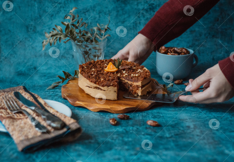 Скачать Женские руки берут лопаточкой кусок домашнего торта с шоколадом и кремом рядом с тарелкой с вилкой и ножом и кружкой, наполненной орехами на темном фоне. Домашняя выпечка фотосток Ozero