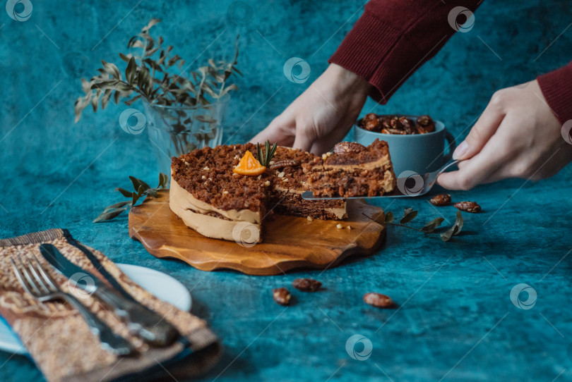 Скачать Женские руки берут лопаточкой кусок домашнего торта с шоколадом и кремом рядом с тарелкой с вилкой и ножом и кружкой, наполненной орехами на темном фоне. Домашняя выпечка фотосток Ozero