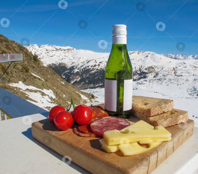 Скачать Бутылка вина, домашняя колбаса, сыр, хлеб и помидоры черри на старой деревянной разделочной доске на фоне заснеженных гор и ярко-голубого неба. Содержание путешествия фотосток Ozero