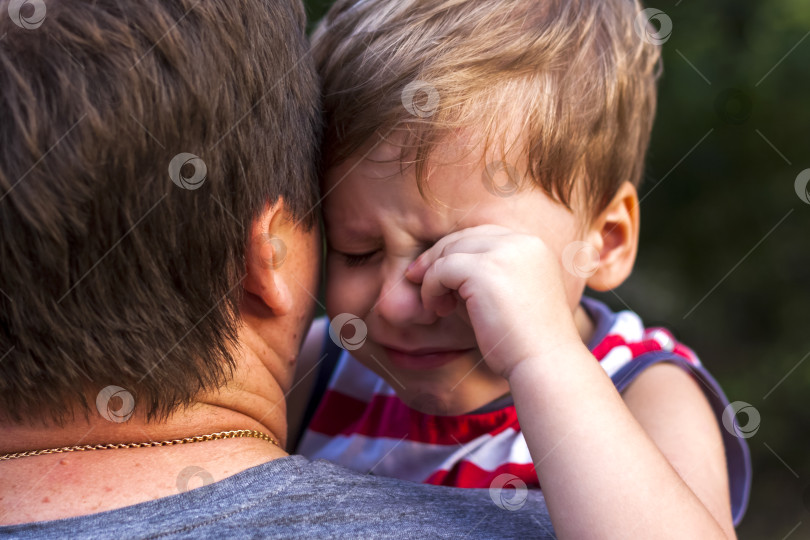 Скачать Маленький мальчик плачет, находясь на руках у своего отца, прижимаясь к нему, чтобы чувствовать себя защищенным. фотосток Ozero
