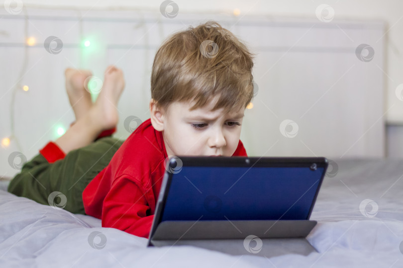 Скачать Милый мальчик сидит на кровати и смотрит в планшет. Забавный улыбающийся ребенок. фотосток Ozero