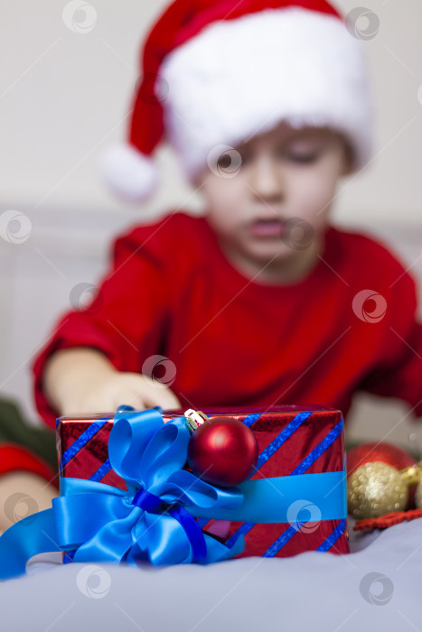 Скачать Рождественский подарок с голубым бантом лежит на фоне симпатичного мальчика в шапке Санта-Клауса. Выборочный фокус. концепция Рождества и Нового года. фотосток Ozero