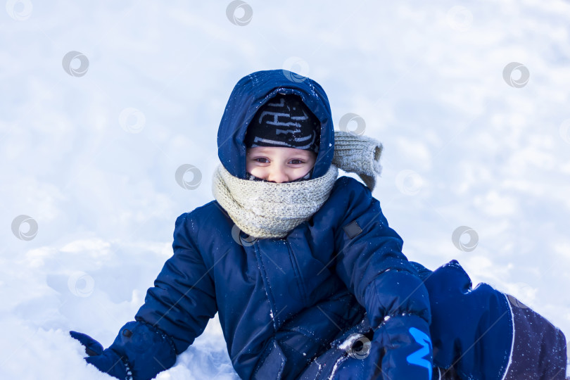 Скачать Улыбающийся счастливый ребенок лежит в сугробе солнечным зимним днем. Много снега и очень морозно. Активные зимние игры на свежем воздухе. фотосток Ozero
