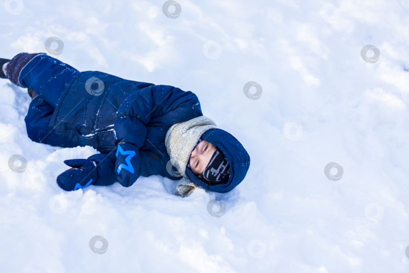 Скачать Улыбающийся счастливый ребенок лежит в сугробе солнечным зимним днем. Много снега и очень морозно. Активные зимние игры на свежем воздухе. фотосток Ozero