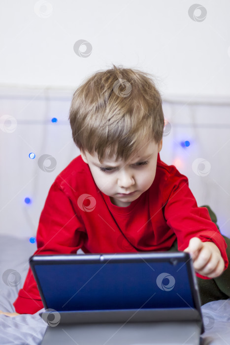 Скачать Симпатичный мальчик лежит на кровати и смотрит в планшет. Онлайн-обучение, общение в Интернете. Забавный улыбающийся ребенок. фотосток Ozero