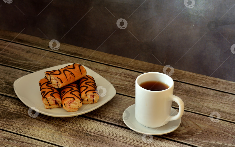 Скачать Тарелка с несколькими мини-бисквитными рулетиками в шоколадной глазури и чашка горячего чая на деревянном столе. фотосток Ozero