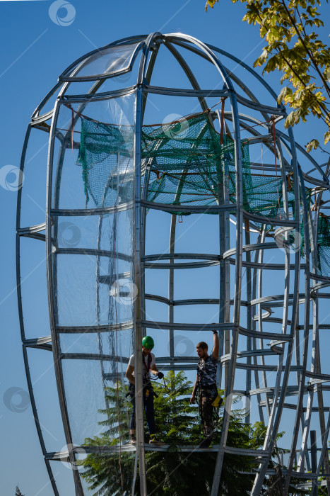 Скачать Рабочие возводят металлические конструкции с прозрачными куполами над вечнозелеными деревьями джакаранды.Фонтан бесконечности.Защита деревьев от ветра и холода. Городской парк Краснодара. фотосток Ozero