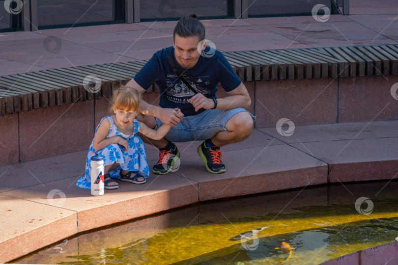 Скачать Молодой папа и его дочь смотрят на рыбок в пруду. Зона отдыха "Зеркальный лабиринт" с деревом Делоникс регия на островке посреди пруда. Общественный городской парк Краснодара. фотосток Ozero