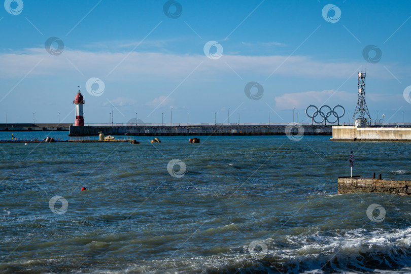 Скачать Действующий городской маяк на Пирсе возле морского вокзала. Голубое небо. Черное море. Справа от маяка олимпийский символ - пять олимпийских колец. Солнечный зимний день. фотосток Ozero