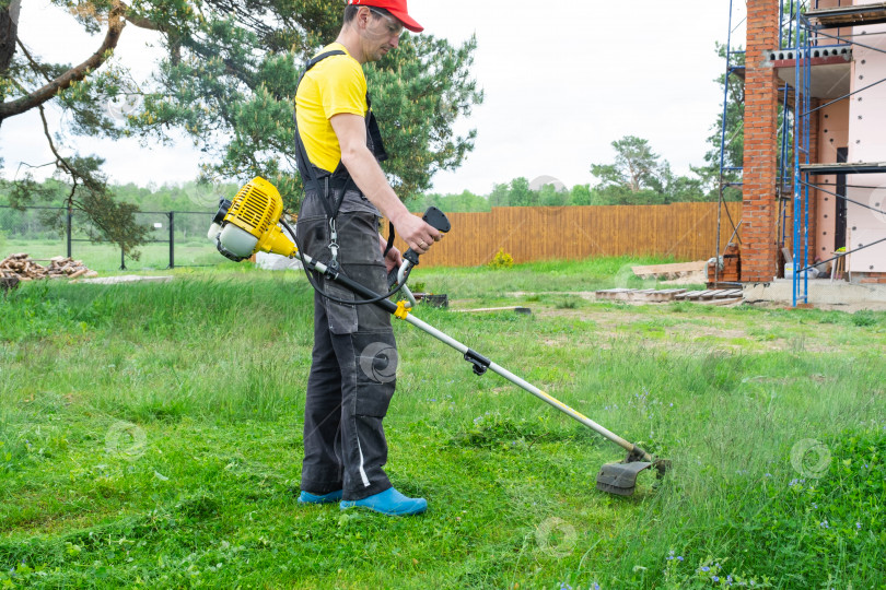Скачать Мужчина-садовник подстригает зеленую траву газона на заднем дворе на строительной площадке бензиновой косилкой. Триммер для ухода за садовым участком фотосток Ozero