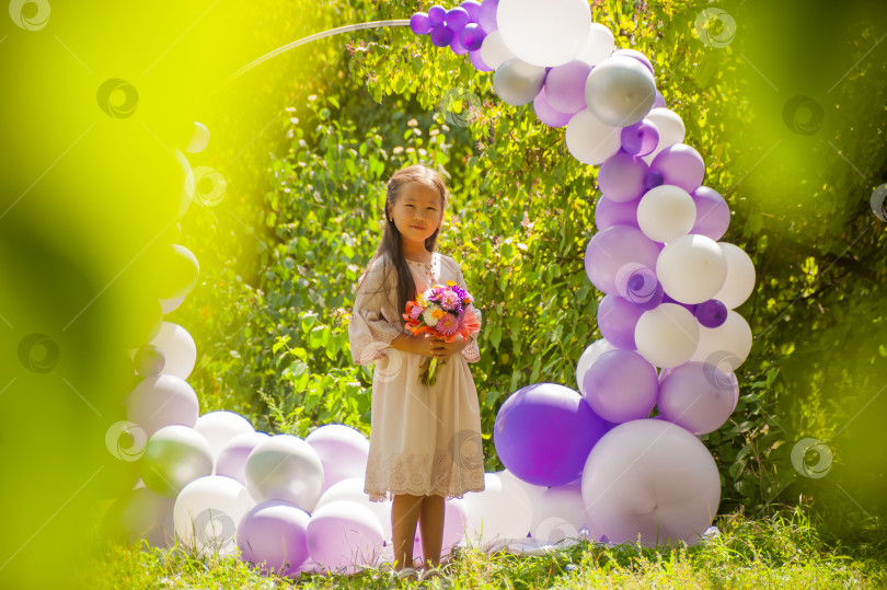 Скачать 7-летняя азиатская девочка позирует в платье среди зелени на фоне арки из воздушных шаров фотосток Ozero