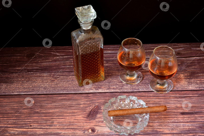 Скачать Кубинская сигара в хрустальной пепельнице, графин и два бокала с коньяком на столе из темного дерева. фотосток Ozero