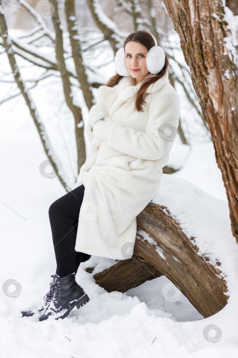 Скачать Счастливая молодая женщина в белой шубе, варежках и меховых наушниках на зимней прогулке в парке фотосток Ozero