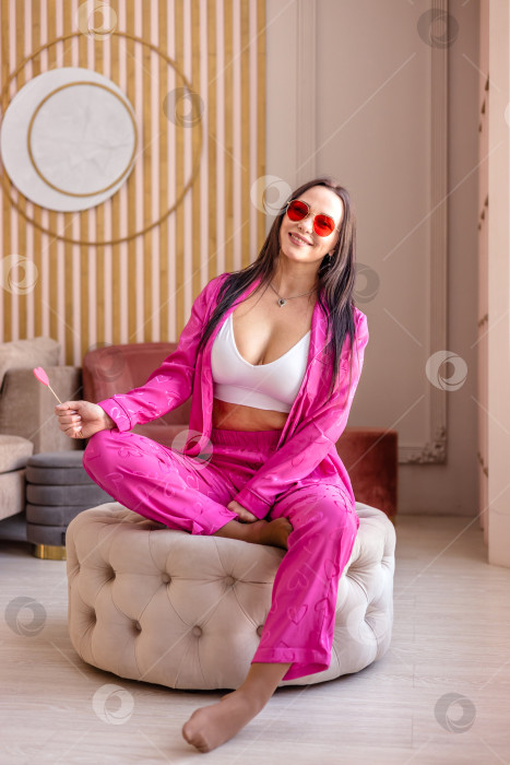 Скачать Красивая молодая брюнетка в розовой пижаме с конфетой на палочке в руках в форме сердца сидит на пуфике в гостиной. день святого Валентина фотосток Ozero