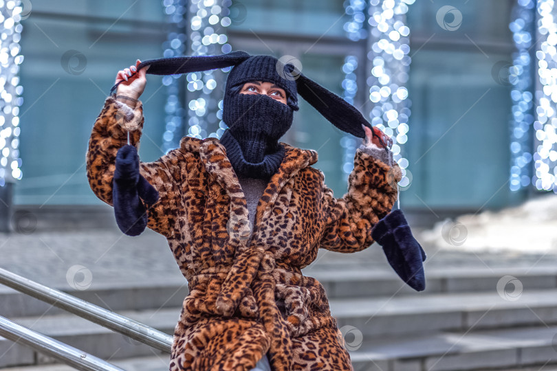 Скачать Стильная молодая женщина в шубе с леопардовым принтом и балаклаве с заячьими ушками на городской улице во время рождественских праздников фотосток Ozero