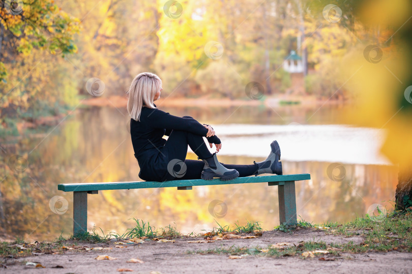 Скачать Привлекательная блондинка спортивного телосложения отдыхает, сидя на скамейке в парке у озера, и смотрит на прекрасный солнечный осенний закат фотосток Ozero
