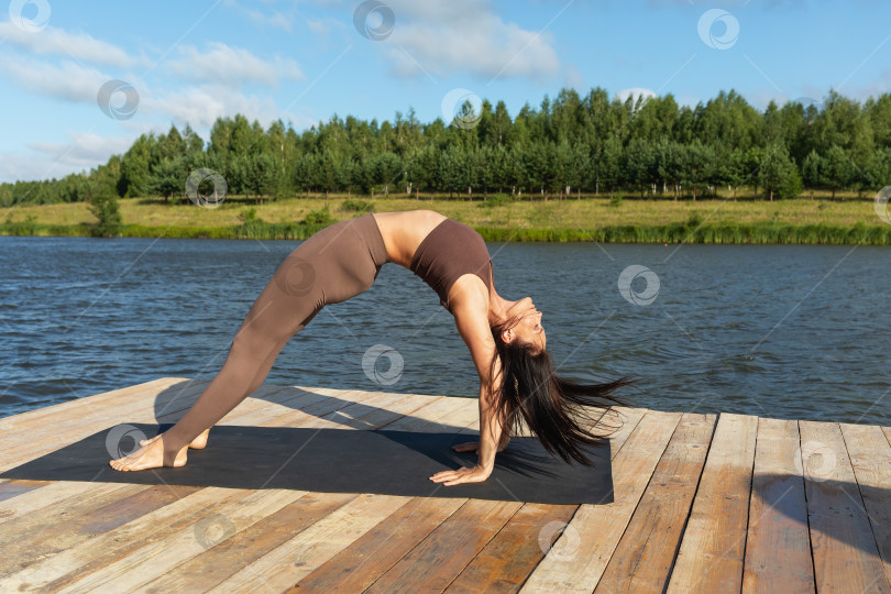 Скачать Привлекательная женщина, выполняющая упражнение Сету Бандхасана, позу моста, тренирующаяся в леггинсах и коротком топе на берегу озера солнечным теплым утром фотосток Ozero