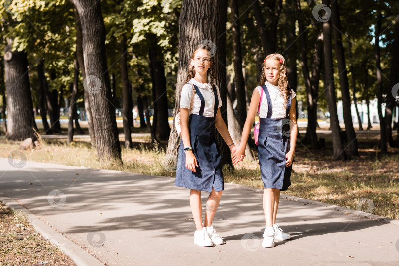 Скачать Две девочки из начальной школы улыбаются, держатся за руки и идут в школу теплым днем фотосток Ozero