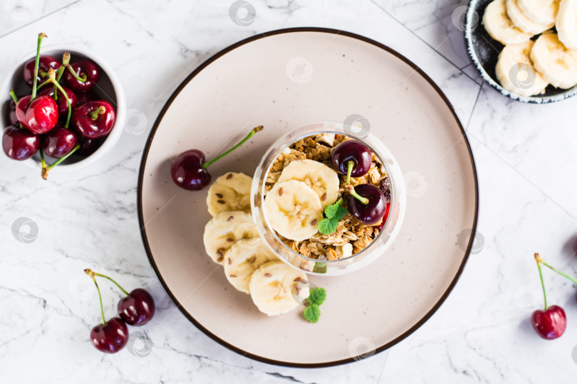 Скачать Гранола с греческим йогуртом, вишней и бананом в стакане на столе. Вид сверху фотосток Ozero