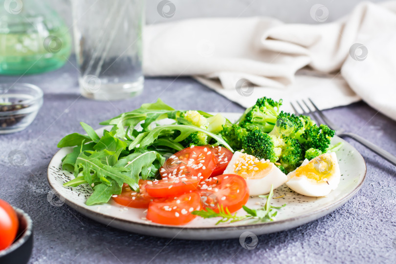 Скачать Тарелка с брокколи, помидорами, рукколой, вареным яйцом и семенами кунжута на тарелке на столе. фотосток Ozero