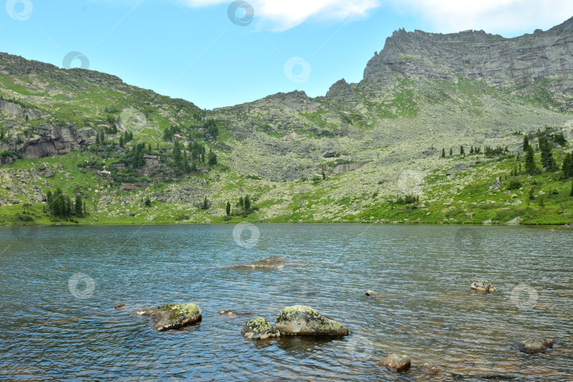 Скачать Плоские камни, торчащие из озера над поверхностью воды, окруженные высокими горными хребтами в пасмурный летний день. фотосток Ozero