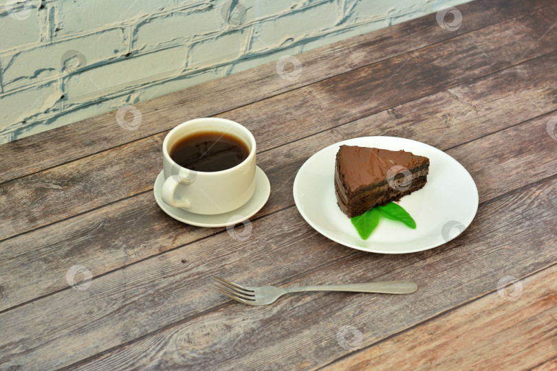 Скачать Тарелка с кусочком шоколадного бисквитного торта с листьями мяты, чашка горячего черного кофе на блюдце и вилка на деревянном столе. фотосток Ozero