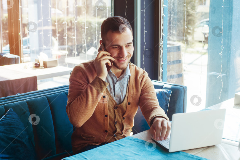 Скачать Молодой человек сидит в кафе, работает за ноутбуком, разговаривает по телефону. Портрет молодого человека. фотосток Ozero