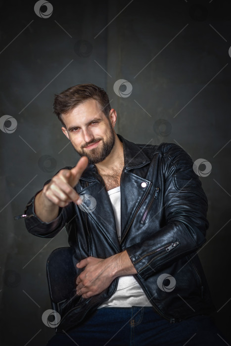 Скачать Современный молодой человек со стильной прической, одетый в классную черную кожаную куртку и сидящий на стуле на темном фоне фотосток Ozero