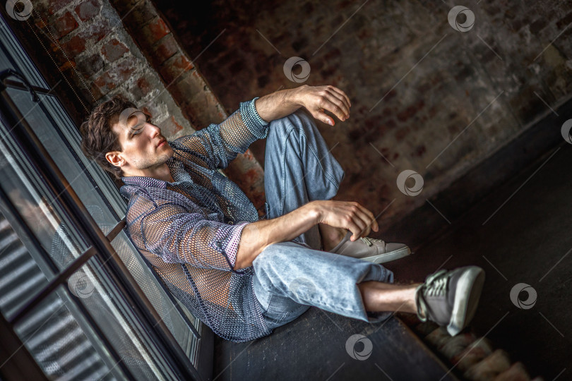 Скачать Современный молодой человек со стильной прической сидит на подоконнике у окна на темном фоне. фотосток Ozero