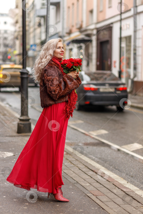 Скачать Элегантная блондинка в красном вечернем платье и шубе стоит на улицах города, ожидая такси. Весенняя концепция, международный женский день. 8 марта фотосток Ozero