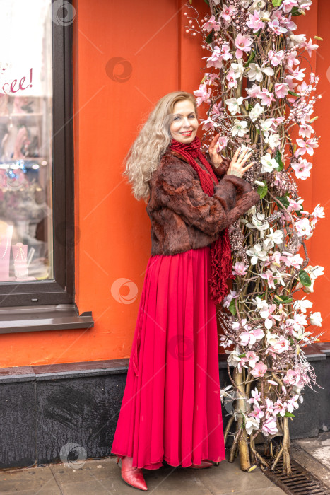 Скачать Элегантная блондинка в красном вечернем платье и шубе на улицах города возле декоративной стены, украшенной цветами магнолии. Весенняя концепция, международный женский день. 8 марта фотосток Ozero