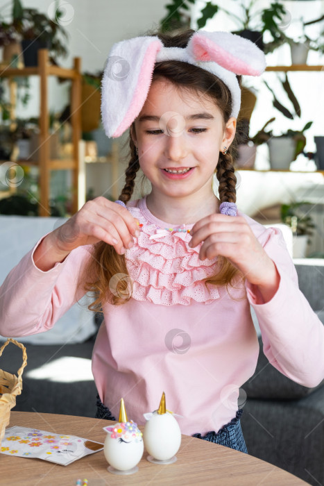 Скачать Милая девочка с розовыми ушками кролика делает пасхальную поделку - украшает яйцо в виде единорога стразами, рогом, цветами в интерьере домика с растениями. фотосток Ozero