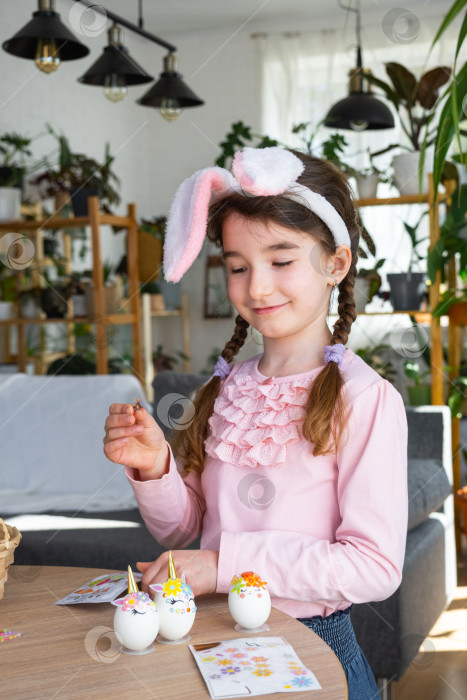 Скачать Милая девочка с розовыми ушками кролика делает пасхальную поделку - украшает яйцо в виде единорога стразами, рогом, цветами в интерьере домика с растениями. фотосток Ozero