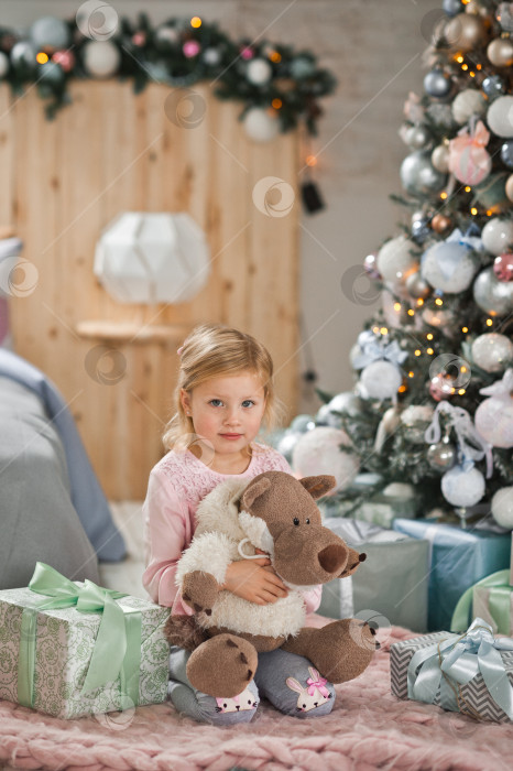 Скачать Счастливый ребенок, обнимающий игрушку, сидит рядом с рождественской елкой 2110. фотосток Ozero