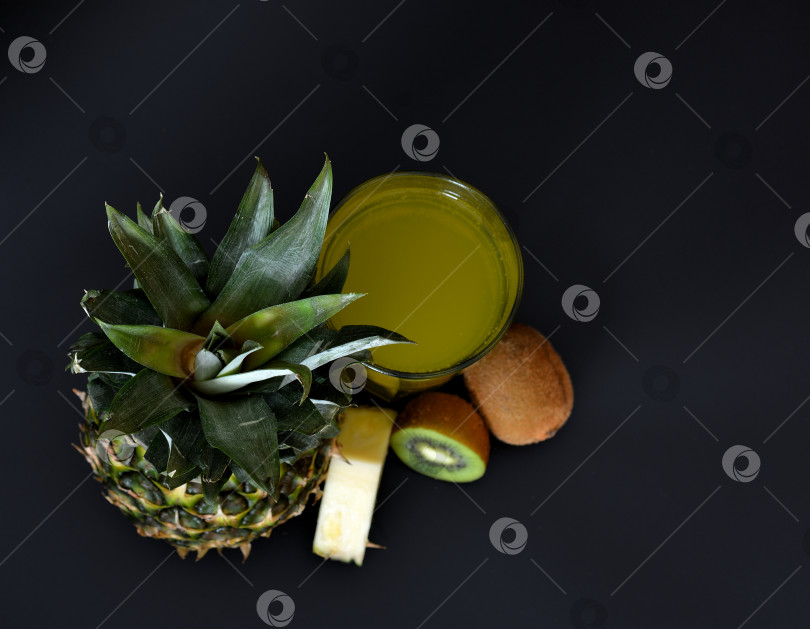 Скачать Стакан сока из тропических фруктов на черном фоне, рядом с кусочками спелого ананаса и киви. фотосток Ozero