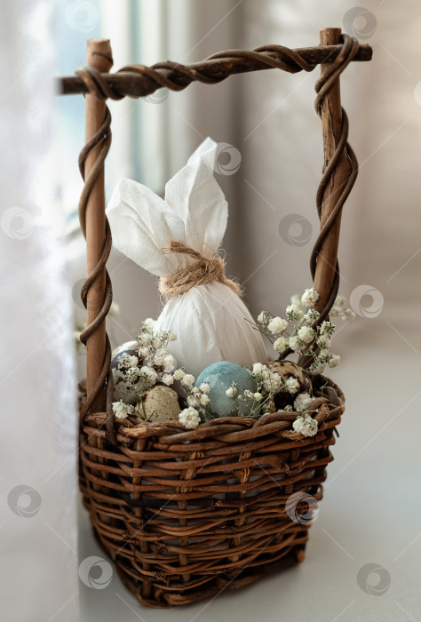 Скачать Пасхальный декор из яйца, украшенного кроличьими ушками, в плетеной корзинке с яйцами и белыми цветами при естественном освещении фотосток Ozero