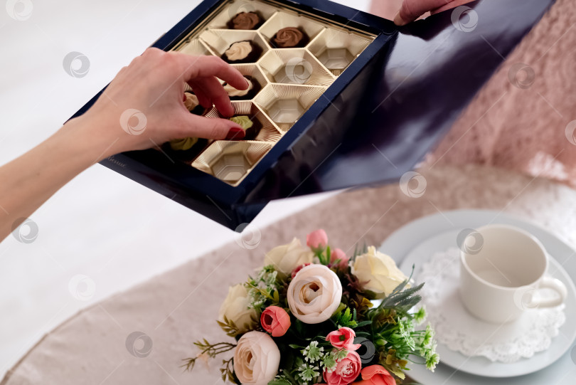 Скачать женская рука берет шоколадную конфету из коробки, протянутой над сервированным журнальным столиком фотосток Ozero
