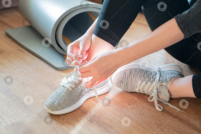 Скачать Женщина сидит на полу рядом со скрученным ковриком и завязывает шнурки на кроссовках, готовясь к занятию йогой фотосток Ozero