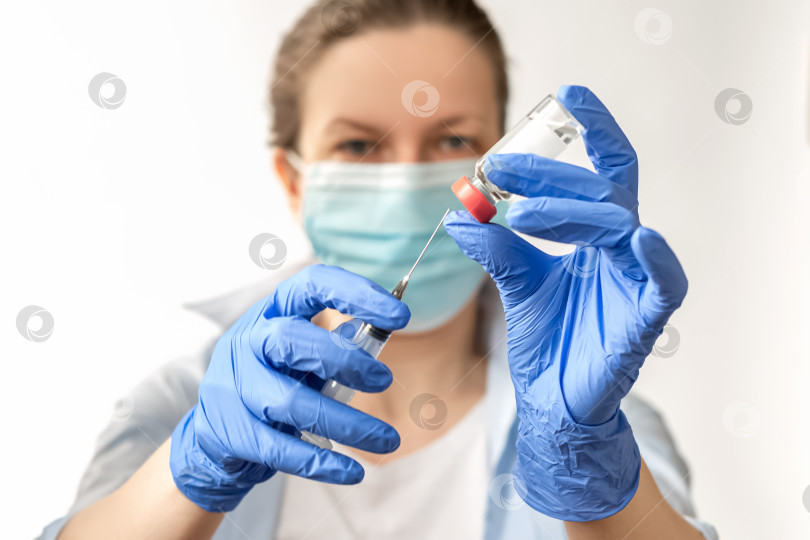 Скачать Женщина в медицинском костюме и защитной маске для лица, в синих латексных перчатках держит ампулу с вакциной covid-19 и шприц, готовый к инъекции. фотосток Ozero