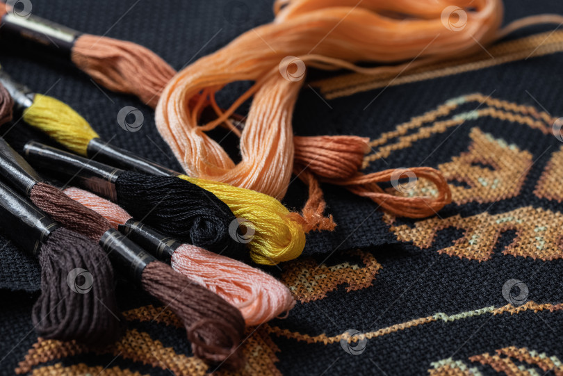 Скачать Разноцветные мотки с нитками мулине разбросаны по черному полотну, покрытому рисунком для вышивания крестиком. фотосток Ozero