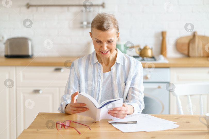 Скачать Счастливая пенсионерка сидит на кухне и наслаждается чтением книги фотосток Ozero